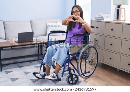 Young hispanic woman sitting on wheelchair at home smiling in love doing heart symbol shape with hands. romantic concept. 