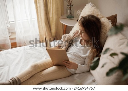 Portrait of happy child. Little beautiful curly girl hugs her plush rabbit and reads book in her bed. Royalty-Free Stock Photo #2416934757