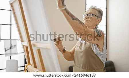 Serious, grey-haired senior woman in apron seriously concentrates on drawing art at indoor studio, embodying her mature creativity on canvas.
