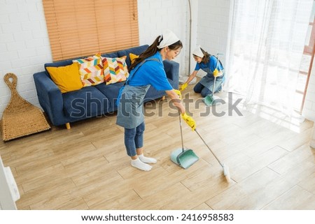 Two Asian young professional cleaning service women worker team working in the house. Girls housekeeper sweeps broomsticks on the wooden floor with another one cleaning under the sofa. Cleaner. Royalty-Free Stock Photo #2416958583