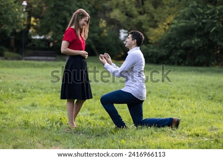 Young man down on his knees, proposing to his girlfriend in the park with an engagement ring.