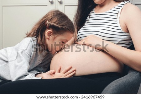 Pregnant woman with her little daughter at home. Cute little girl kisses her mother's tummy. Happy pregnant woman her child, expectation.