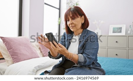 A senior hispanic woman engaging with a smartphone in a cozy bedroom setting, reflecting technology use in later life. Royalty-Free Stock Photo #2416974569