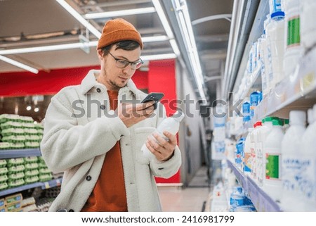 Man in store takes picture of label using phone. Young bearded Caucasian guy in glasses in department with dairy products and eggs holds kefir in hands, takes photo of composition, comparing.