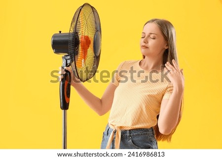 Sweaty young woman with electric fan on yellow background Royalty-Free Stock Photo #2416986813
