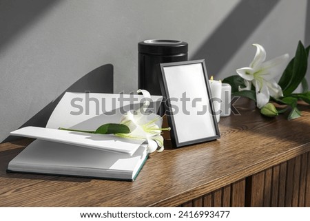 Blank funeral frame, mortuary urn, book and lily flowers on wooden cabinet in room Royalty-Free Stock Photo #2416993477