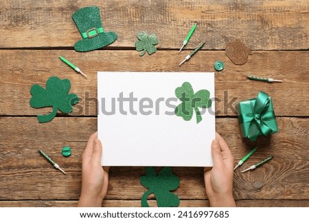 Female hands with blank card, gift box and decor on wooden background. St. Patrick's Day celebration