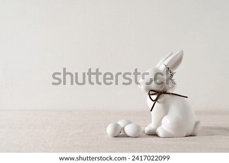Minimalist aesthetic Easter holiday background, white porcelain bunny figurine decor and small candy eggs on light neutral beige linen tablecloth, soft natural sunlight, selective focus.