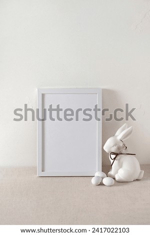 Empty white picture frame or poster mockup, white bunny decorative figurine, small eggs on neutral beige linen table background, Easter holiday business branding template.