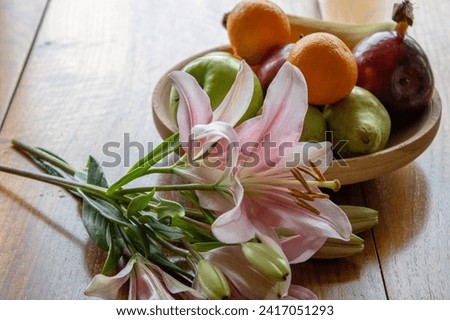 A bunch of flowers laying in front of a wooden fruit bowl standing on a backlit wooden surface with vibrant fruit colors and colorful green and pink peony.