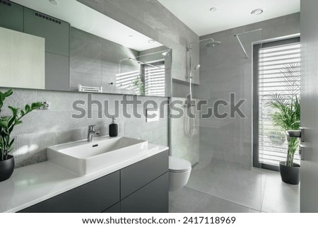 The image shows a modern bathroom with grey tiles, a white sink, mirror, toilet, and a glass shower. There are two green plants for a natural touch. Natural light comes in through a window.