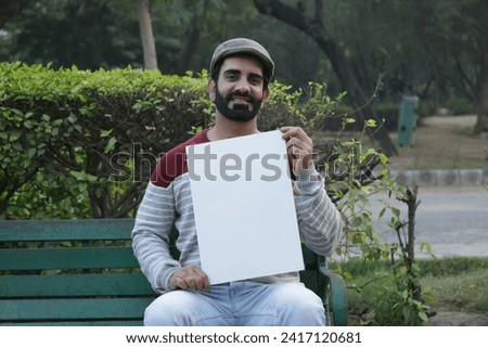 Portrait of attractive bearded young man showing white board and sitting bench in a natural environment. Handsome Indian young man sitting outside looking at camera. Wearing handsome cap.