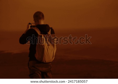 back rear view of one alone backpacker Man with rucksack, cell phone standing on peck of rock, taking photos of the view. Man on top of a mountain photographing sky above Black sea or pacific ocean.