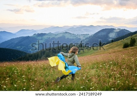 Happy girl running on hills with Ukrainian flag on shoulders. Back view of female child flutting Ukrainian flag. Little kid running in Carpathian mountains. Royalty-Free Stock Photo #2417189249