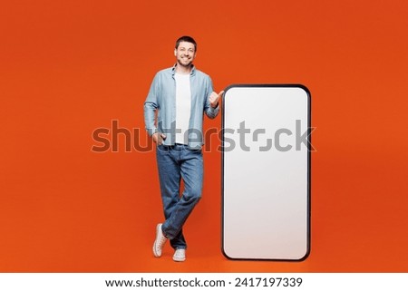 Full body young man wears blue shirt white t-shirt casual clothes point thumb finger on big huge blank screen mobile cell phone smartphone with workspace area isolated on plain red orange background