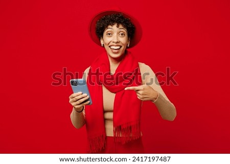 Young happy fun woman of African American ethnicity she wears beige sweater hat scarf hold in hand use point finger on mobile cell phone look camera isolated on plain red background. Lifestyle concept