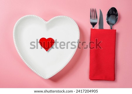 A heart plate with a red heart and silverware of fork, knife, and spoon in a red napkin on a pink background. Romantic dinner Valentine's Day concept