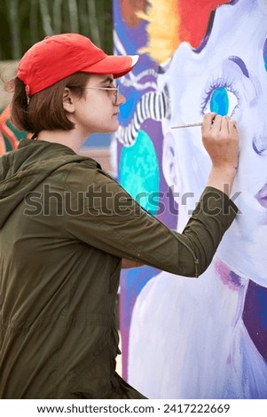 Female painter in red cap draws surreal picture with paintbrush on canvas for outdoor street exhibition, close up side view of female artist apply brushstrokes to canvas, symphony of art creativity