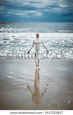 Hairless girl with alopecia in white futuristic suit comes out of cold restless sea on sandy beach, metaphoric performance of bald female artist about overcoming challenges of life and self acceptance