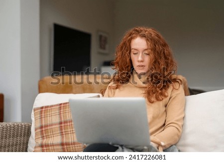 Happy young redhead woman in brown shirt and using laptop, studying, learning, shopping, working online, while sitting on a couch at home