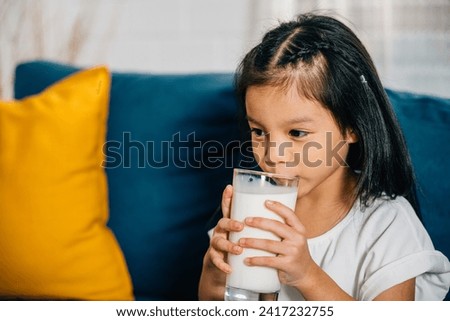 Portrait of a cheerful Asian child savoring a glass of milk on the sofa radiating happiness and innocence. A heartwarming image symbolizing significance of nourishing kids with calcium-rich goodness.