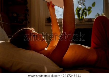 Cute child, boy, lying in bed with book, reading in the morning, sunny weather wintertime