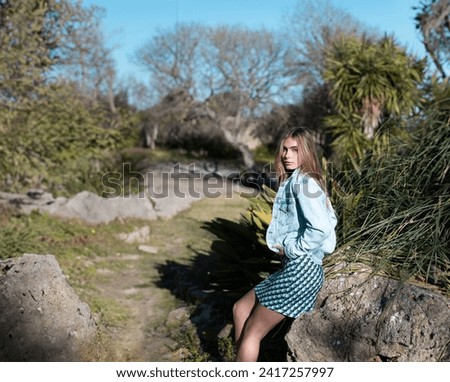 Nature surrounded by plants: Young blond girl enjoying the sun in her face, surrounded by green plants and trees, wearing a nice short patterned dress and a jean jacket - Lifestyle on a sunny day 