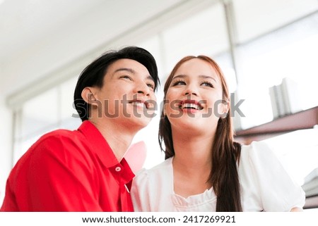 A young happy Asian couple looking up together, sitting, embracing and smiling on Valentine's Day at home. Asian woman enjoys her anniversary date with her boyfriend. Valentine's Day celebration.
