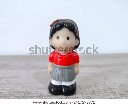 A plastic doll of a girl wearing a red t-shirt and gray skirt standing on a white background