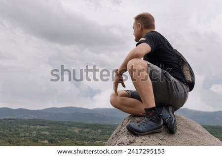Young man sitting on a rock at the top of a mountain peak looking at the beautiful views on a cloudy day. Brave man achieves success. Freedom and contemplation concept. Inspiration and motivation