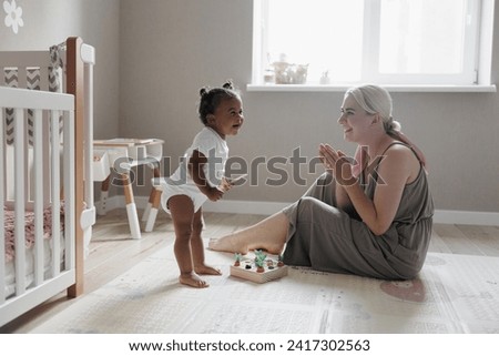 Joyful caucasian mother sits on floor in nursery and plays with her laughing biracial toddler daughter. Concept of interracial family.