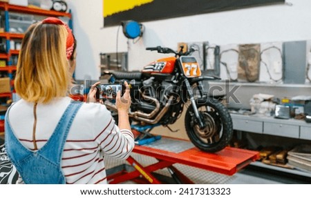 Unrecognizable female mechanic taking photo with cellphone to custom motorcycle repaired over platform on garage. Selective focus on phone screen.