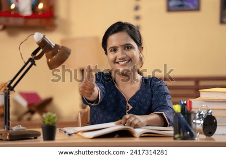 Happy Indian girl showing thumbs up while preparing for examination or studying by looking camera - concept of ambition, educational development and aspirant. Royalty-Free Stock Photo #2417314281