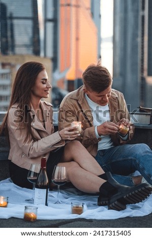 Young loving couple on a surprise date on rooftop on Saint Valentine's Day. Laughing, kissing embracing, drinking wine, having candlelit picnic with cityscape urban view with skyscrapers on background