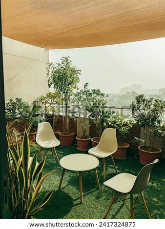 Image shows cozy balcony garden with various potted plants creating lush greenery. The space is furnished with modern white chairs and a table. inviting visitors to relax and enjoy the natural beauty.