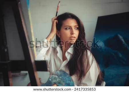 Pensive lovely drawing woman artist with brush painting oil paints on canvas, thought looking away. Perfect lady painter in white shirt sit in art lab. Artisanal lifestyle concept. Copy ad text space