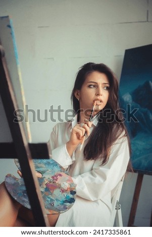 Perfect lady painter in white shirt painting oil paints on canvas, thought looking away. Pensive lovely drawing woman artist with brush sit in art lab. Artisanal lifestyle concept. Copy ad text space