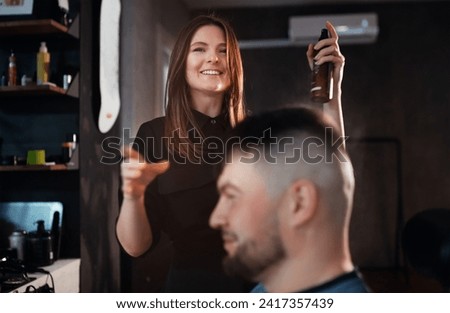 Smiling Young female hairdresser spraying eau de toilette perfume after bearded man hairstyling. Modern low light black style barber shop interior. Hair care service, local small business concept imag Royalty-Free Stock Photo #2417357439