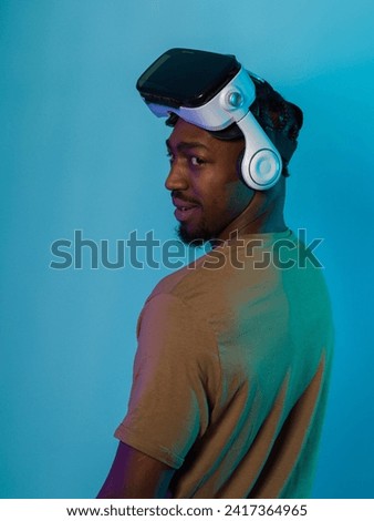 In a futuristic visual, an African American man stands isolated against a striking blue backdrop, adorned with VR glasses that transport him into a cutting-edge virtual reality experience, merging