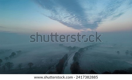 This evocative aerial image captures the tranquil beauty of dawn as it breaks through a layer of mist over a frosty winter landscape. The sky, filled with a dramatic formation of clouds, gradually Royalty-Free Stock Photo #2417382885