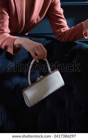 Silver metallic sparkling purse held by woman hands on a blue velour armchair background. Creative shiny woman hand bag studio photography. Royalty-Free Stock Photo #2417386297