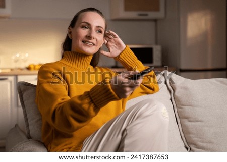 A smiling woman lounges comfortably on a sofa, holding a remote control for an evening of entertainment in her inviting, warm-lit living space. Royalty-Free Stock Photo #2417387653