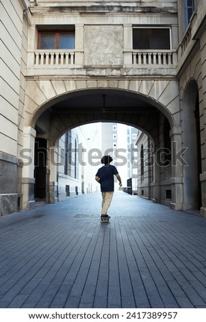Rear view of unrecognizable teen boy on skateboard riding under the arch of building.