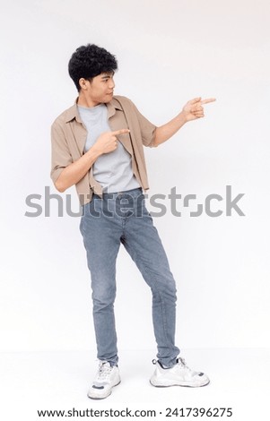 A happy young Asian man with curly hair pointing to the side, smiling and standing against a white studio background. Royalty-Free Stock Photo #2417396275