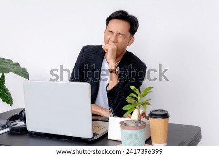 Young Asian man at desk with laptop yawning, showing signs of boredom. At a modern office setting. Royalty-Free Stock Photo #2417396399