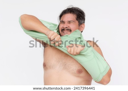 An overweight man struggles to put on an undersized polo shirt past his chest. A shirt too small or consequence of gaining too much weight. Isolated on a white background. Royalty-Free Stock Photo #2417396441