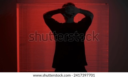 Portrait of male in dark room. Handsome man near window, red neon light shines behind jalousie, guy facing window holding hand over head.