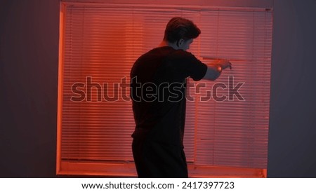 Portrait of male in dark room. Handsome man near window, red neon light shines behind jalousie, guy looks outside tries to open blinds with finger.