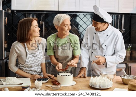 joyous young woman in apron smiling at chef and her mature friend that decorating cake with berries Royalty-Free Stock Photo #2417408121