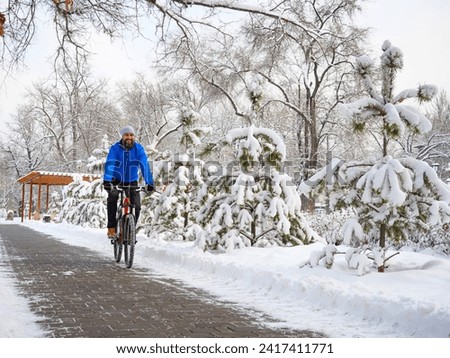 A joyful man rides a bicycle in a winter park among snow-covered trees. Bearded cyclist in a blue jacket.. Active lifestyle